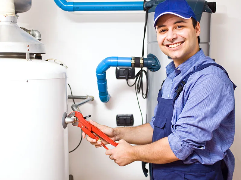 A man in overalls stands beside a water heater, holding a wrench, ready for maintenance or repair work.