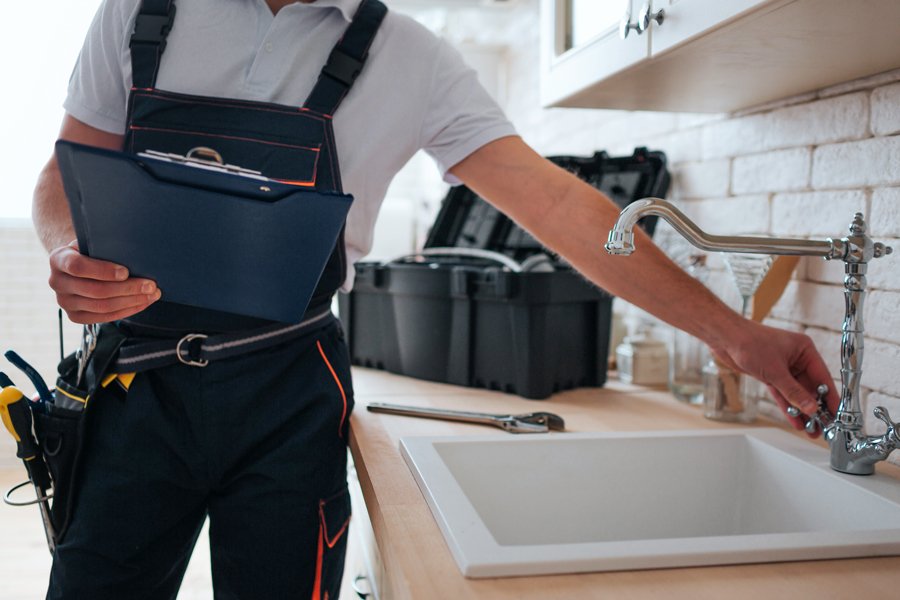 A plumber stands beside a sink, holding a clipboard, assessing the plumbing situation with a focused expression.